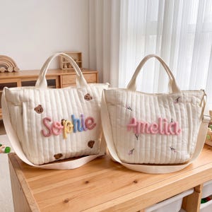 May include: Two off-white quilted tote bags with shoulder straps. The left bag features the name Sophie in multi-colored yarn lettering and small embroidered bears. The right bag displays the name Amelia in pink yarn with floral embroidery.