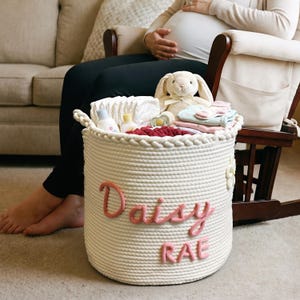 May include: A white rope basket filled with baby items, including a stuffed bunny, diapers, and baby clothes. The basket has the name "Daisy Rae" in pink letters on the side. The basket is sitting on a carpeted floor.