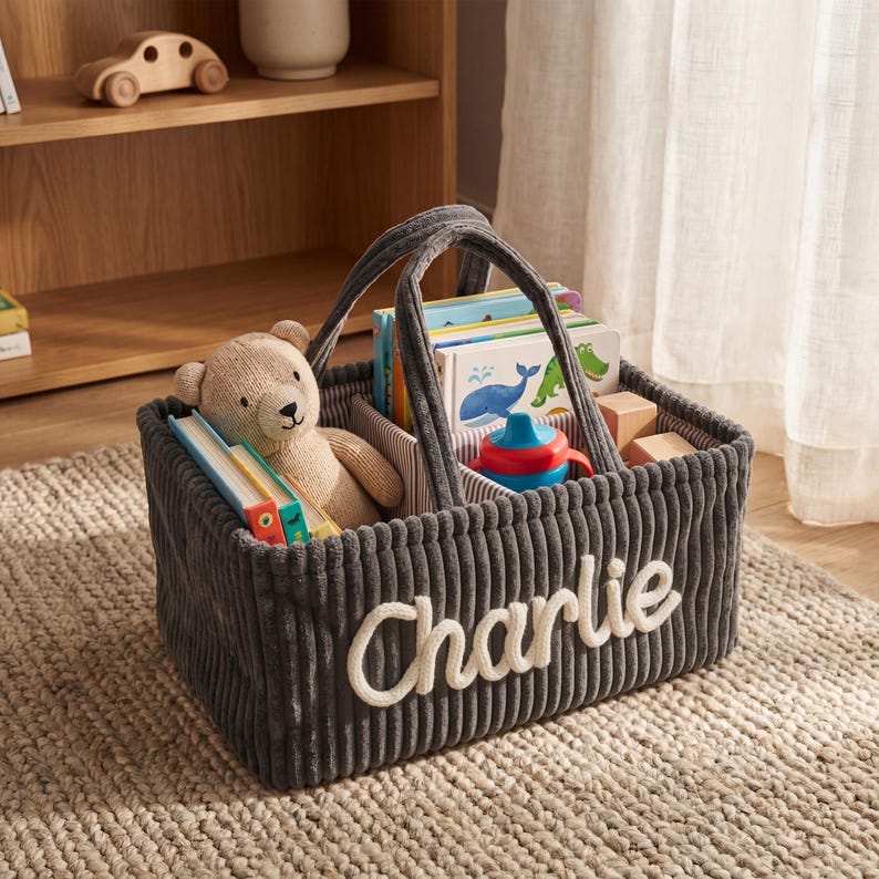May include: A gray corduroy storage basket with the name "Charlie" embroidered in white. The basket contains books, a teddy bear, and a sippy cup. The basket has a handle for easy carrying.