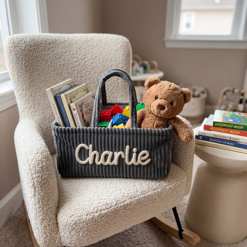 May include: A gray corduroy tote bag with the name "Charlie" embroidered in white, filled with books, colorful blocks, and a teddy bear. The tote is on a cream-colored armchair, suggesting a children's room setting.