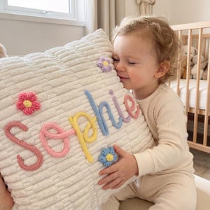 Peut inclure: Un coussin en velours côtelé blanc avec le nom "Sophie" en lettres tricotées colorées et plusieurs motifs de fleurs tricotées. Le coussin est tenu par un enfant.