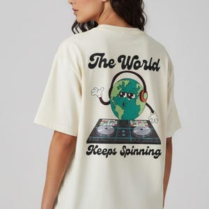 May include: An off-white short-sleeve t-shirt featuring a graphic on the back. The design shows a cartoon Earth wearing headphones and sunglasses, standing behind a DJ turntable console. The text reads The World Keeps Spinning in black font.