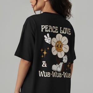 May include: A black short-sleeved t-shirt featuring a graphic on the back. The design includes a cartoon flower character with a face, white petals, and brown centre, making a peace sign. Text reads PEACE LOVE & WUB-WUB-WUB with small gold star accents.