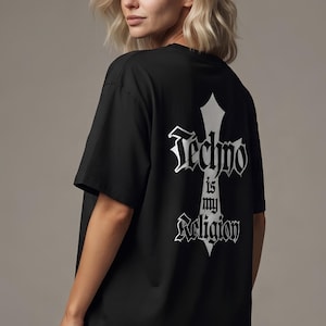 May include: A black short-sleeved t-shirt featuring a white graphic on the back. The design includes a vertical cross shape with the text Techno is my Religion written in a gothic-style font centered over the cross graphic on the fabric.