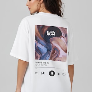 May include: A white short-sleeve t-shirt featuring a large rectangular graphic on the back. The graphic displays an abstract swirl of blue, brown, and cream colors with the text RÜFÜS DU SOL, Innerbloom, and a digital music player interface.