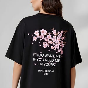May include: Black t-shirt with a floral design of pink cherry blossoms and the text "IF YOU WANT ME, IF YOU NEED ME, I'M YOURS". The shirt also has the text "INNERBLOOM 5:46".