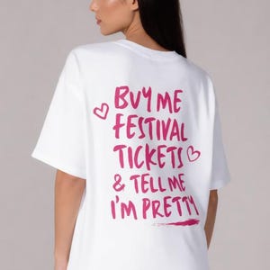 May include: A white short-sleeved t-shirt featuring pink text on the back that reads "BUY ME FESTIVAL TICKETS & TELL ME I'M PRETTY" accompanied by two small pink heart outlines. The shirt has a crew neckline and a loose, relaxed fit.