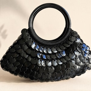May include: A black handbag covered in layered, circular, reflective sequins. The bag features a rigid, circular black handle attached to the top, creating a distinct crescent-shaped silhouette. The surface is entirely textured with overlapping black discs.