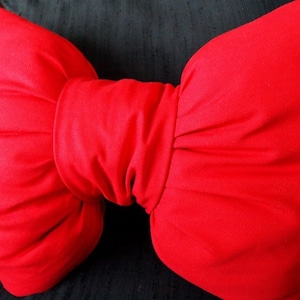 Peut inclure: Un nœud papillon rouge vif en tissu, avec un centre froncé et des extrémités arrondies. Le nœud est placé sur un fond sombre, mettant en valeur sa couleur vibrante et sa forme. Le tissu semble lisse et finement tissé.