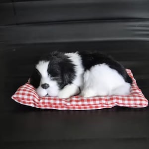 May include: A plush, sleeping dog figurine with black and white fur, resting on a red and white checkered pillow. The dog is lying down, appearing relaxed. The pillow is rectangular.