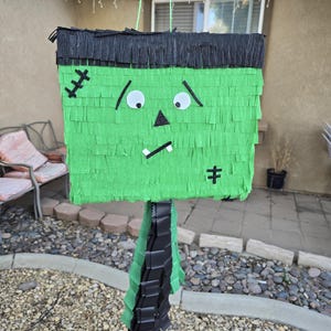 May include: A green Frankenstein's monster piñata with black hair and facial features. The piñata is made of layered paper and has a rectangular head and a fringed body. It is hanging from a green string, perfect for a Halloween party.
