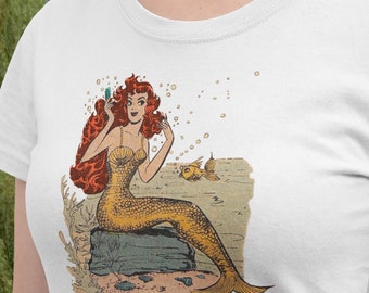 May include: A white t-shirt with a graphic of a red-haired mermaid holding a mirror and looking at her reflection. The mermaid is wearing a seashell bra and has a gold tail. The graphic is surrounded by bubbles.