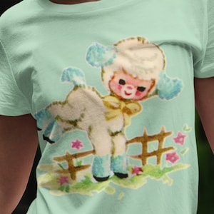 Peut inclure: T-shirt vert clair avec une illustration r&eacute;tro d'un agneau sautant par-dessus une cl&ocirc;ture en bois. L'agneau a une fourrure blanche duveteuse, des d&eacute;tails bleus et un n&oelig;ud jaune. Des fleurs roses et de l'herbe verte sont &eacute;galement visibles.
