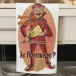May include: A white kitchen towel with a cartoon devil holding a block of cheese. The devil is wearing a red suit and has horns and a beard. The text on the towel reads "Avez-vous essayé le fromage?"