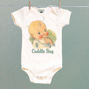 Puede incluir: Un body blanco para bebé con una ilustración vintage de un bebé con cabello rubio y ojos azules. El bebé lleva una camisa de lunares azules y blancos y mira hacia un lado con un dedo en la boca. El texto "Cuddle Bug" está impreso en turquesa debajo de la ilustración.