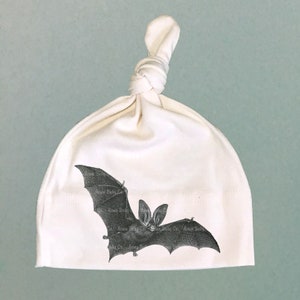 May include: A white knotted baby hat with a black and white illustration of a bat.