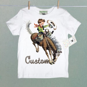 K&ouml;nnte beinhalten: Wei&szlig;es T-Shirt mit einem Motiv eines Jungen, der ein wildes Pferd reitet. Der Junge tr&auml;gt einen Cowboyhut und ein rotes Bandana. Der Text "Custom" ist in braunen Seilliteren unter dem Motiv geschrieben.