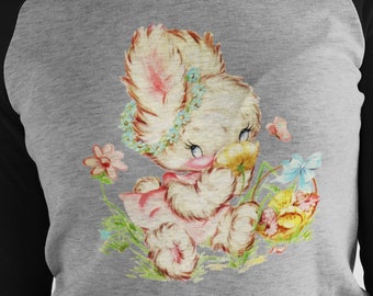 May include: A grey and black baseball tee shirt with a vintage illustration of a white bunny rabbit wearing a flower crown and holding a yellow flower. The bunny is surrounded by flowers and butterflies.