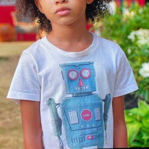 Puede incluir: Una persona joven con cabello casta&ntilde;o rizado usando una camiseta blanca con un gr&aacute;fico de un robot azul. El robot tiene ojos rojos y una nariz roja.