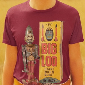 May include: A person wearing a maroon t-shirt with a vintage toy robot printed on the front. The robot is holding a red laser gun and is standing in front of a yellow box with the words "Big Loo Giant Moon Robot by Marx" printed on it.