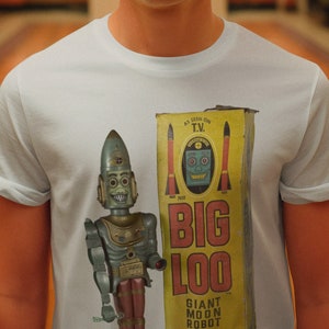 May include: A man wearing a white sailor hat and a light blue t-shirt with a vintage toy robot printed on the front. The robot is holding a gun and is standing in front of a yellow box with the words "Big Loo Giant Moon Robot" printed on it.
