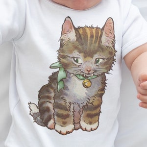 Peut inclure: Body bébé blanc avec une illustration vintage d'un chat tigré portant un ruban vert et une cloche. Le chat est marron, gris et beige. Le bébé porte un bandeau blanc.