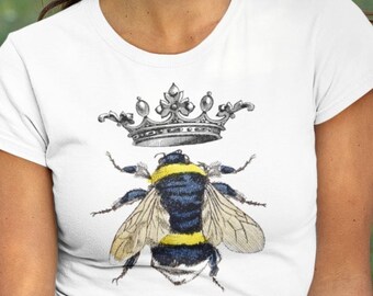 Peut inclure: T-shirt blanc avec un motif d'abeille portant une couronne. L'abeille est noire et jaune avec des ailes blanches. La couronne est argentée.