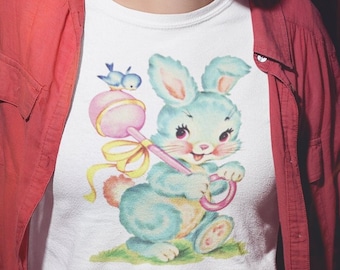 May include: A white t-shirt with a vintage illustration of a blue bunny holding a rattle and a ring. The bunny is surrounded by yellow and pink accents.