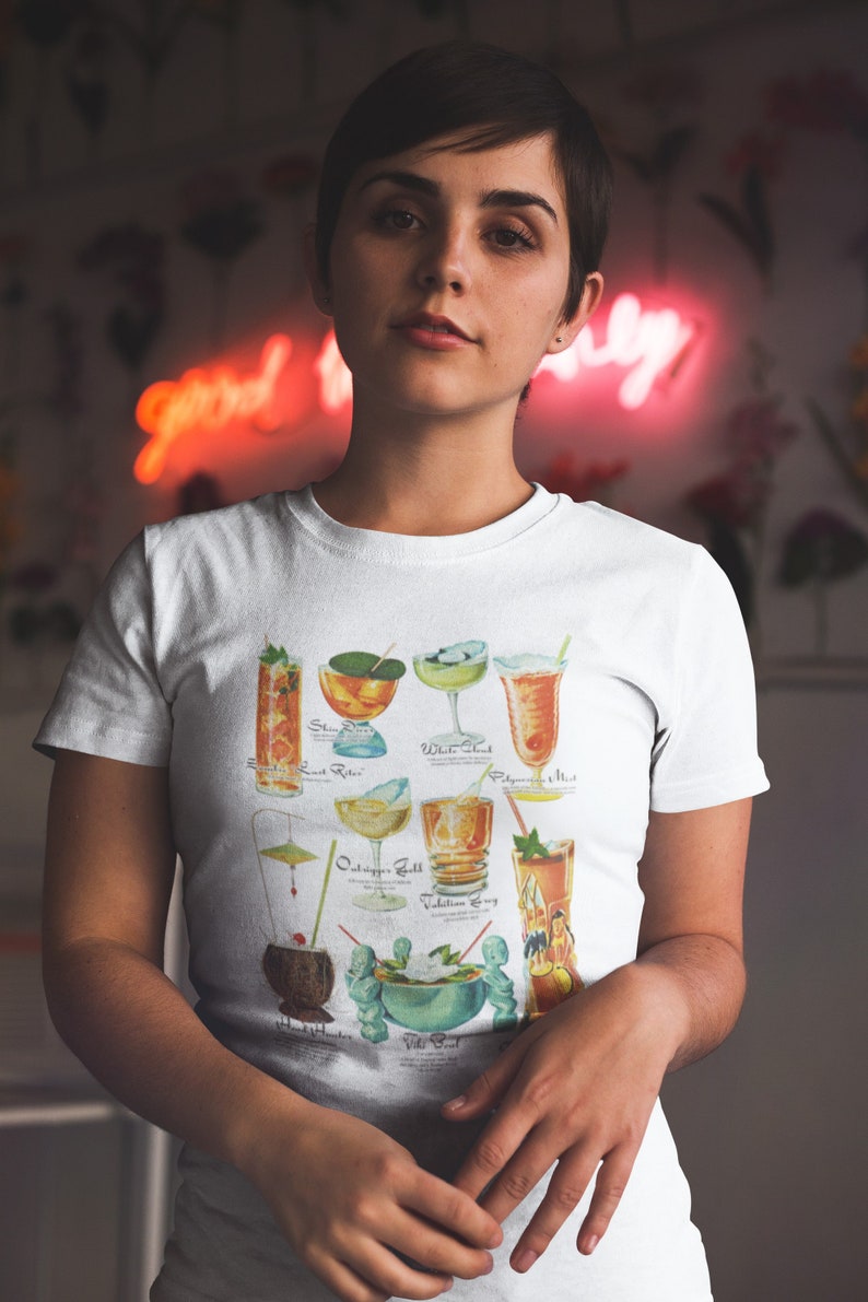 Op de afbeelding: Wit T-shirt met een vintage illustratie van verschillende cocktails. De cocktails zijn gelabeld met hun namen: "Singapore Sling", "Last Rider", "White Shark", "Polynesian Mist", "Outrigger Zest", "Vodka Zing", "Island Hummer", "Vili Vili".