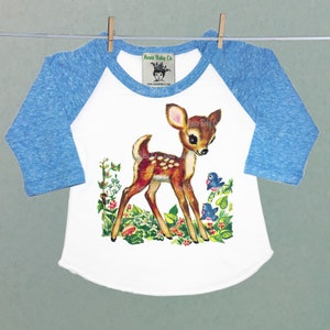 May include: A white baby onesie with blue raglan sleeves features a vintage illustration of a brown fawn standing in a field of green leaves and red flowers with two blue birds. The text "Acme Baby Co." is printed on the onesie.