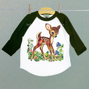 May include: White, green, and brown baseball tee shirt with a vintage illustration of a fawn with two bluebirds in a field of flowers. The text "Acme Baby Co." is printed on the shirt.