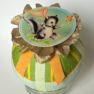 May include: A decorative egg-shaped ornament with a green, orange, and blue striped pattern. The top features a circular image of a cartoon skunk chasing a butterfly with a net. Gold-colored paper frills surround the image.