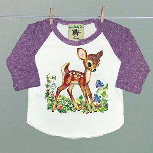 May include: A white and purple baby onesie with a vintage illustration of a fawn in a forest setting. The onesie has a raglan sleeve design and the text "Acoma Baby Co." on the front.