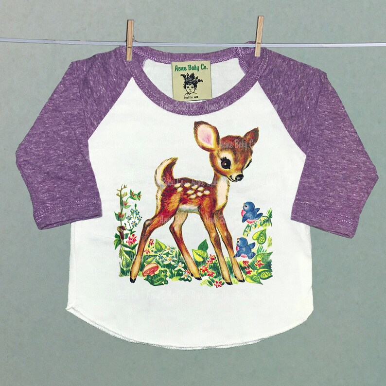 May include: A white and purple baby onesie with a vintage illustration of a fawn in a forest setting. The onesie has a raglan sleeve design and the text "Acoma Baby Co." on the front.