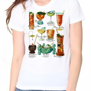 Op de afbeelding: Wit T-shirt met een kleurrijke illustratie van verschillende tropische cocktails. De namen van de cocktails staan ​​onder elke afbeelding. De cocktails omvatten: Zombie, Last Rites, Shin-Dig, White Cloud, Polynesian Mist, Outrigger Gold, Valhallian Swing, Head Hunter, Tiki Bowl en Foggy Gutter.