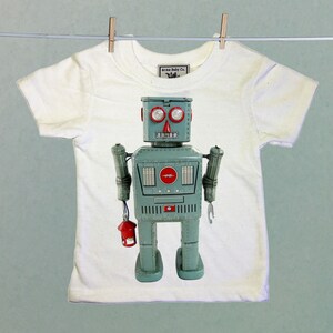 Puede incluir: Una camiseta blanca con un robot vintage impreso en la parte delantera. El robot es azul y gris con detalles rojos. El robot tiene una luz roja en el pecho y una linterna roja en la mano. La camiseta tiene una peque&ntilde;a etiqueta que dice "Acme Baby Co."