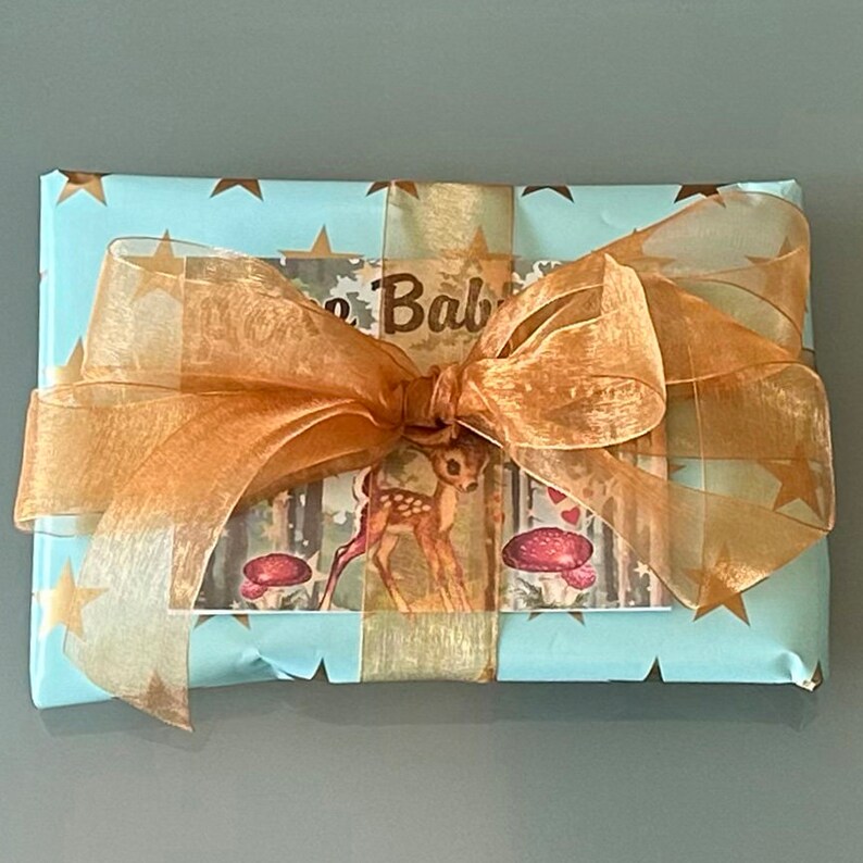 Op de afbeelding: Een lichtblauwe cadeaudoos met gouden sterren en een gouden lint gebonden in een strik. De doos heeft een kaart bovenop met een afbeelding van een hert in het bos en de tekst "The Baby is Here".