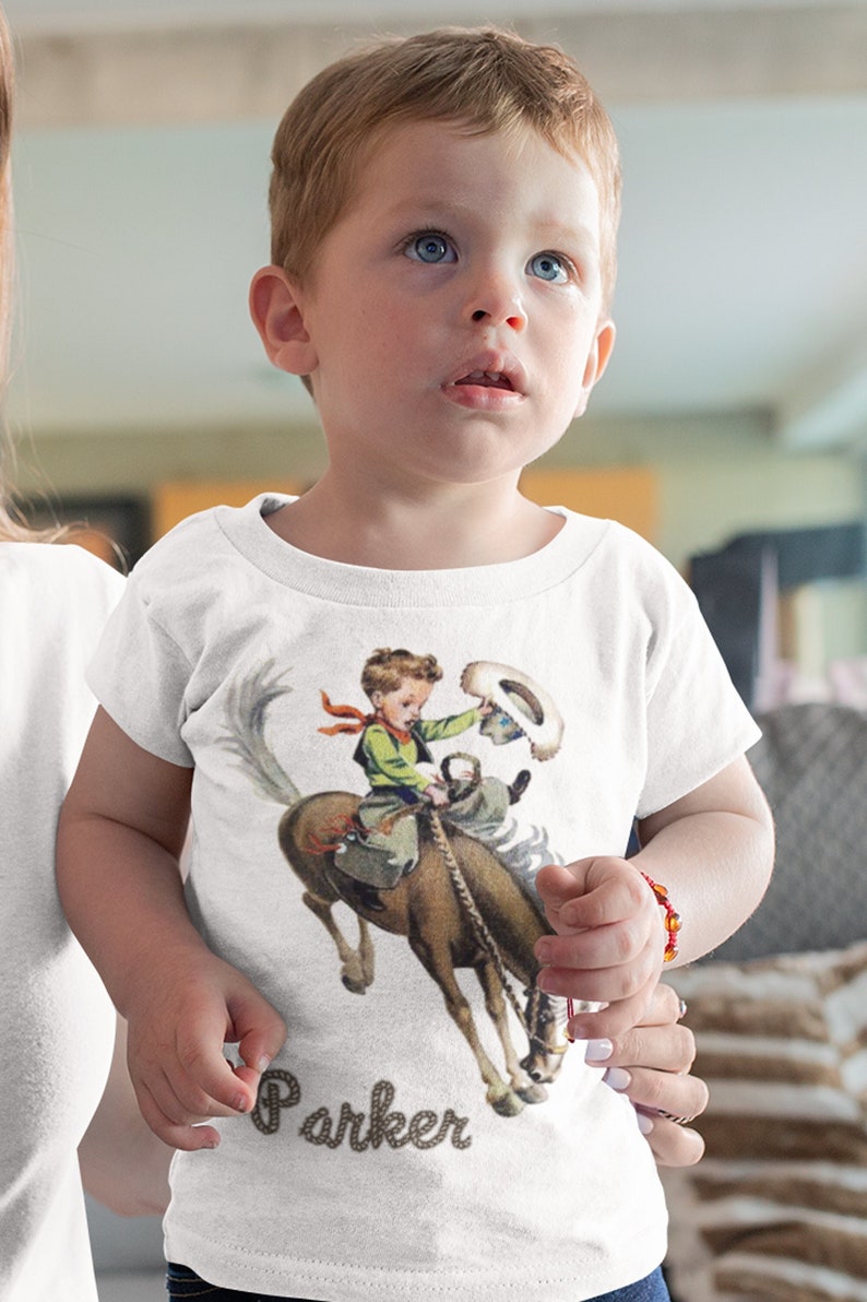 K&ouml;nnte beinhalten: Wei&szlig;es T-Shirt mit einem Aufdruck eines Jungen, der ein sich aufb&auml;umendes Pferd reitet. Der Text "Parker" ist auf das T-Shirt gedruckt.