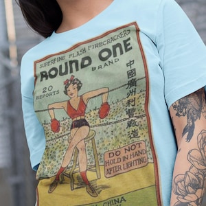 Puede incluir: Camiseta azul claro con un diseño vintage de fuegos artificiales "Round One". El diseño incluye una ilustración retro de una mujer con atuendo de boxeo, con texto en inglés y chino. Fabricado en China.