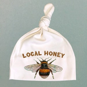 May include: White knotted baby hat with the text "LOCAL HONEY" in brown lettering and a graphic of a bumblebee.