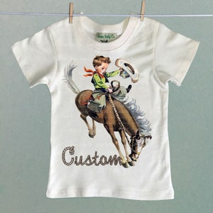 K&ouml;nnte beinhalten: Ein wei&szlig;es T-Shirt mit einer Vintage-Illustration eines Jungen, der ein sich aufb&auml;umendes Pferd reitet. Der Text "Custom" ist in braunen, seilartigen Buchstaben unter dem Bild gedruckt.