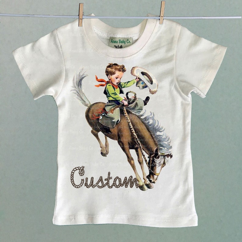 K&ouml;nnte beinhalten: Ein wei&szlig;es T-Shirt mit einer Vintage-Illustration eines Jungen, der ein sich aufb&auml;umendes Pferd reitet. Der Text "Custom" ist in braunen, seilartigen Buchstaben unter dem Bild gedruckt.