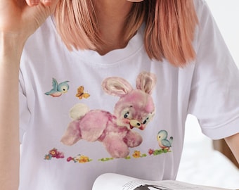 May include: White t-shirt with a pink bunny rabbit graphic. The bunny is running and has a blue bird flying next to it. There are also butterflies and flowers on the shirt.