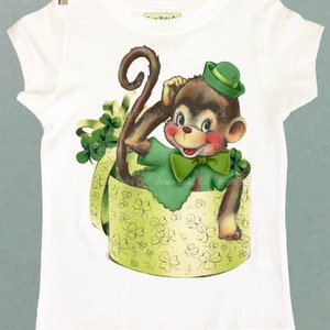 May include: A white t-shirt with a green shamrock print of a monkey wearing a green hat and holding a shamrock. The monkey is sitting in a green box with shamrock print.