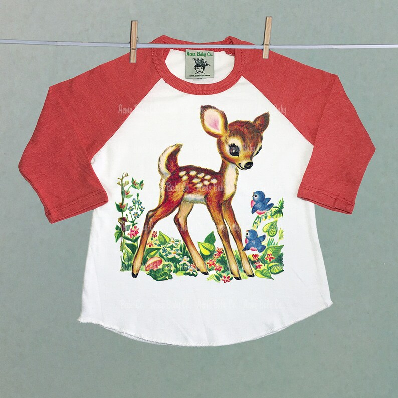 May include: A white and orange baseball tee shirt with a vintage illustration of a fawn with two bluebirds. The text "Acme Baby Co." is printed on the shirt.