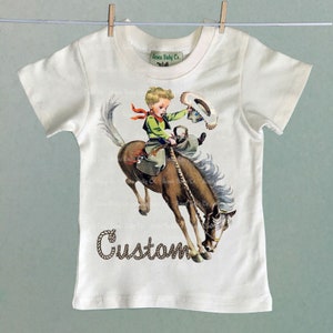 K&ouml;nnte beinhalten: Wei&szlig;es T-Shirt mit einer Vintage-Illustration eines Jungen, der ein sich aufb&auml;umendes Pferd reitet. Der Junge tr&auml;gt ein gr&uuml;nes Hemd, eine braune Hose und ein rotes Bandana. Das Pferd ist braun mit einer wei&szlig;en M&auml;hne und einem wei&szlig;en Schweif. Der Text "Custom" ist in braunem Seil unter dem Bild geschrieben.