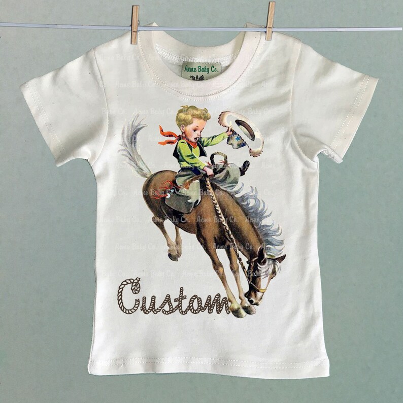 K&ouml;nnte beinhalten: Wei&szlig;es T-Shirt mit einer Vintage-Illustration eines Jungen, der ein sich aufb&auml;umendes Pferd reitet. Der Junge tr&auml;gt ein gr&uuml;nes Hemd, eine braune Hose und ein rotes Bandana. Das Pferd ist braun mit einer wei&szlig;en M&auml;hne und einem wei&szlig;en Schweif. Der Text "Custom" ist in braunem Seil unter dem Bild geschrieben.
