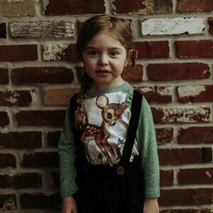 May include: A young girl wearing a green long-sleeved shirt with a white deer graphic and a black corduroy skirt with suspenders. She is standing in front of a brick wall.