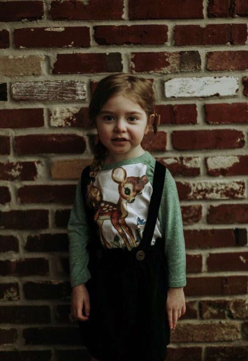 May include: A young girl wearing a green long-sleeved shirt with a white deer graphic and a black corduroy skirt with suspenders. She is standing in front of a brick wall.