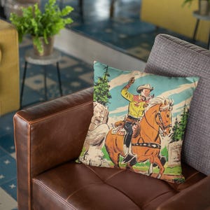 May include: A brown leather couch with a throw pillow featuring a vintage illustration of a cowboy riding a horse through a forest. The pillow has a light blue, green, and yellow color scheme.